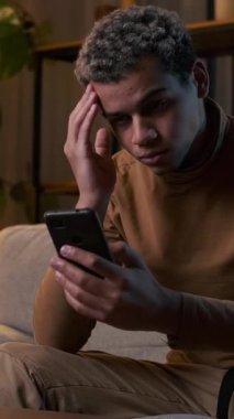 Karanlık bir odada kanepede otururken akıllı telefonuyla kötü haber alan endişeli bir adamın dikey videosu..