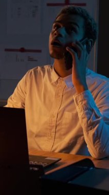 Karanlık bir ofiste geç saatlere kadar çalışan ve önemli bir telefon görüşmesine cevap veren genç bir iş adamının dikey videosu..