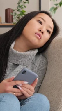 Evdeki kanepede cep telefonunu tutarken kendini yalnız ve sıkılmış hisseden üzgün bir Asyalı kadının dikey videosu..