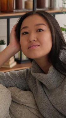 Evdeki kanepede dinlenen genç ve güzel Asyalı bir kadının dikey videosu. Gülümsüyor ve kameraya bakıyor..