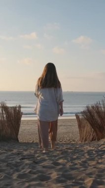 Güzel bir gün batımında kumlu bir yolda çıplak ayakla okyanusa doğru yürüyen bir kadının dikey videosu..