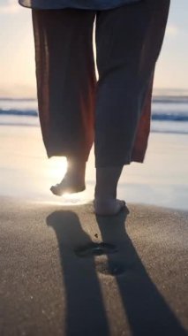 Gün batımında ıslak kumsalda çıplak ayakla yürüyen bir kadının dikey videosu. Kamera, o yumuşak okyanus dalgalarına adım atarken onu takip ediyor..