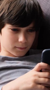 Uzanmış akıllı telefon kullanan bir çocuğun dikey videosu. Evde odaklanmış ve meşgul görünürken ekrana hafifçe vuruyor..