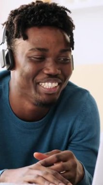 Kulaklıklı mutlu bir Afro-Amerikan adam evdeki bilgisayarındaki bir video görüşmesi sırasında konuşuyor ve gülüyor. Dikey video.