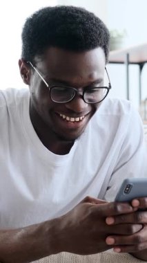 Gözlüklü, gülümseyen ve gülen genç bir Afro-Amerikan adamın evde akıllı telefonunu kullanırken çekilmiş dikey videosu..