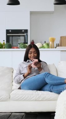 Genç siyahi bir kadın akıllı telefonunu kullanarak gülümsüyor, modern dairesinde bir kanepede oturuyor. Dikey video.