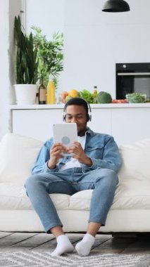 Kulaklıklı genç bir Afrikalı Amerikalı evdeki bir kanepede dinlenirken dijital tablet kullanıyor. Dikey video.