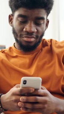 Genç siyahi adam cep telefonunda geziniyor ve mesajlaşıyor, evde otururken farklı duygular gösteriyor. Dikey video.