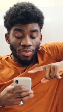 Afro saç stili tomarları olan genç bir zenci evde dinlenirken cep telefonunun içeriğine tepki veriyor. Dikey video.