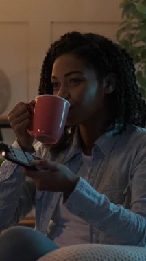 Geceleri televizyon seyreden, kupadan su içen ve oturma odasındaki kanepede otururken uzaktan kumanda kullanan mutlu bir kadın. Dikey video.