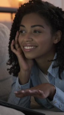 Kıvırcık saçlı neşeli genç bir kadın dizüstü bilgisayar kullanırken gülümsüyor, sıcak bir oturma odasında kanepede uzanıyor. Dikey video.