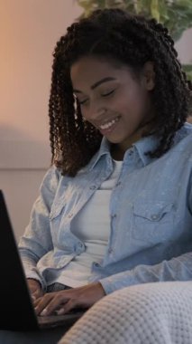 Evde dizüstü bilgisayarla çalışan mutlu bir Afro-Amerikan kadının dikey videosu. O rahat bir koltukta oturuyor, gülümsüyor ve klavyede yazıyor..