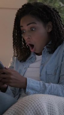 Akşamları kanepede otururken akıllı telefonundan iyi haberler alan şaşırmış bir kadının dikey videosu..