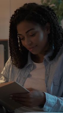 Geceleri rahat bir odada kitap okuyan mutlu genç bir kadının dikey videosu. Kitaptan başını kaldırıp kameraya gülümsüyor..