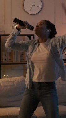 Mutlu kadın uzaktan kumandaya şarkı söylüyor ve geceleri oturma odasında dans ediyor. Tasasız ev eğlencesi ve eğlence. Dikey video.