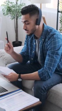 Kulaklıklı genç adam, evde çalışırken bilgisayarındaki bir video görüşmesi sırasında belgeleri tartışıyor. Dikey video.