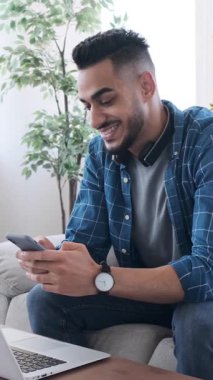 Genç bir adam kanepede oturuyor, akıllı telefonuyla sosyal medyada mesajlaşırken ya da gezinirken yürekten gülüyor. Dikey video.