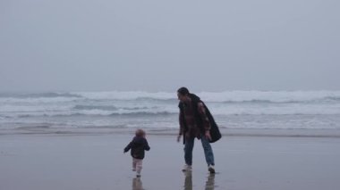 Genç bir anne ve küçük kızı serin ve bulutlu bir günde kumlu bir plajda oynayıp eğleniyorlar. Kadın çocuğu alır ve döndürür..
