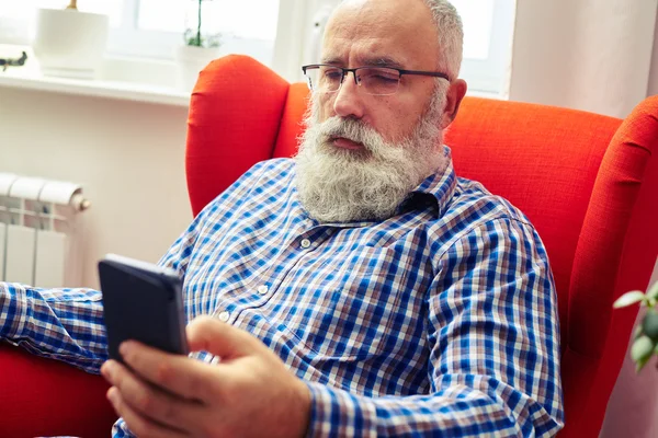 Starší muž na židli s smartphone doma — Stock fotografie