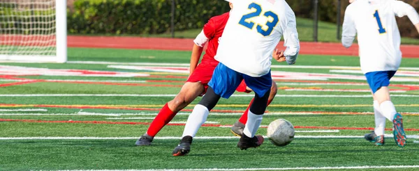 Rear View of a soccer player in a white jersey dribbling the ball around his opponent in a red jersey during a high school game on a green turf field.