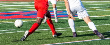 Beyazlı, kırmızı üniformalı liseli futbolcular çim sahasındaki bir maçta topu kovalıyorlar..