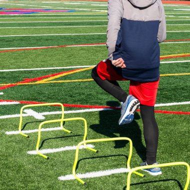 Lise atletizm koşucusunun arka görüntüsü, buz gibi bir bahar günü sahada atletizm antrenmanında bir ayak üzerinde mini engelleri aşmaktır..