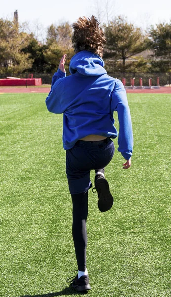 A rear view of a high school runner practicing speed and agility sports drills on a green turf field with his hair bouncing up and down.
