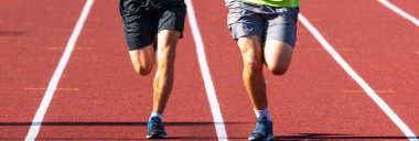 Parlak gün ışığında kırmızı bir pistte yan yana koşan iki koşucunun bacakları ön tarafta..