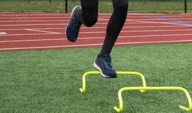 Bir erkek koşucunun, bir çim tarlasında sarı mini engellerin üzerinden atlarken ön görüntüsü..