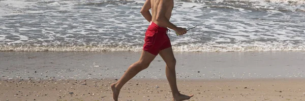 Side view of one male lifeguard running shirtless on the beach in red shorts close to the water