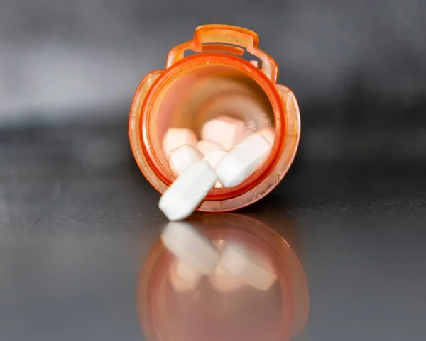 Close up of an opened orange prescription pill bottle full with white pills reflecting on a black counter top.