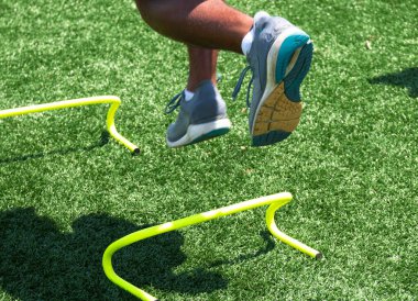 Liseli çocuk yeşil bir çim sahasında sarı mini hurdaların üzerinden atlarken trakc ve saha antrenmanı sırasında.