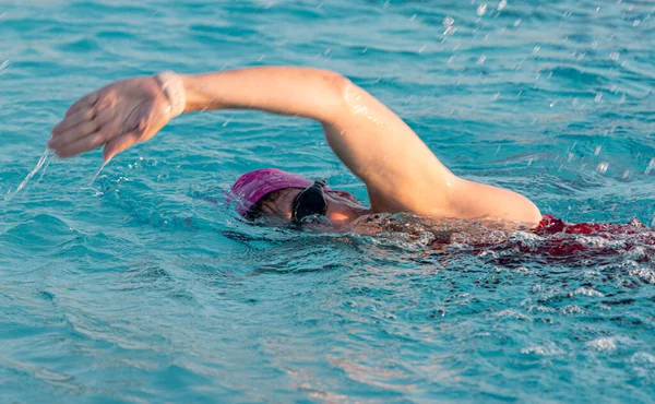 Close up of a female swiming in a pool with her arm raised in the air swimming freestyle wearing a pink cap and googles.