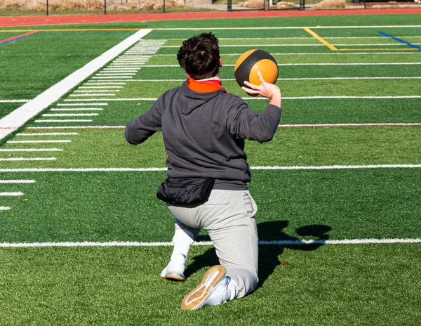 Rear view of a shot put track and field athlete on his knees training by throwing a medicine ball on a green turf field.