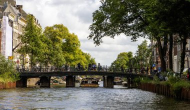 Amsterdam, Hollanda - 5 Ağustos 2025: Ziyaretçiler köprüden geçerken tekneler güneşli gökyüzünün altında süzülüyor.