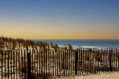 Sahil manzarası, su kenarında ahşap bir çit ve uzun otları gösteriyor. Gökyüzü açık..