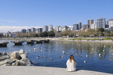 İstanbul, Türkiye - 21 Nisan 2021: Genç bir kadın beton iskelede tek başına oturuyor ve İstanbul 'daki Suadiye Sahili' ni izliyor. Birçok insan plajda dinleniyor ve bahar güneşinin tadını çıkarıyor. Modern binalar görünür..