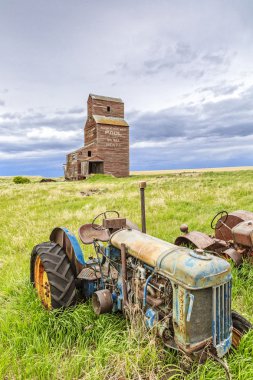 Birkaç eski paslı traktör, Saskatchewan 'ın hayalet kasabası Bents' te terk edilmiş bir tahıl asansörünün altında çayırlarda paslanıyor..