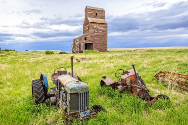 Birkaç eski paslı traktör, Saskatchewan 'ın hayalet kasabası Bents' te terk edilmiş bir tahıl asansörünün altında çayırlarda paslanıyor..