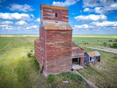 Üzerinde bir sürü odun olan büyük kırmızı tahıl asansörü. Bina eski ve üzerinde çok fazla pas var. Gökyüzü mavi ve bazı bulutlar var.