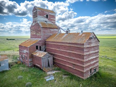 Kırmızı çatılı tahıl asansörü tarlada duruyor. Bina terk edilmiş ve bakımsız görünüyor.