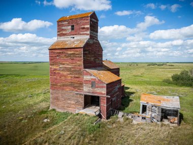 Eğimli çatılı kırmızı tahıl asansörü bir tarlada duruyor. Bina eski ve terk edilmiş.