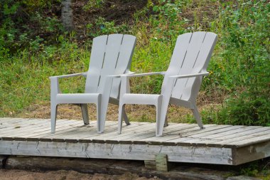 Two chairs are sitting on a wooden platform. The chairs are grey and are facing each other. The platform is surrounded by grass and trees