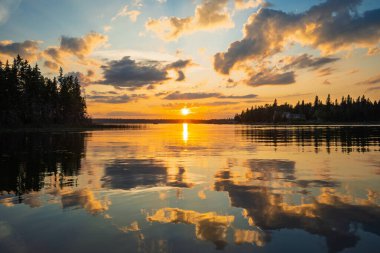 Güneş bir gölün üzerinde batıyor, suyun üzerine sıcak bir parıltı saçıyor. Gökyüzü bulutlarla dolu. Huzurlu ve huzurlu bir atmosfer yaratıyor.