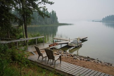 Rıhtımda iki sandalye ve bir tekne var. Rıhtım ağaçlarla çevrili ve su sakin.