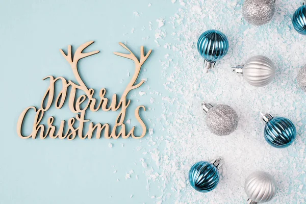 Christmas postcard with wishes concept. Top overhead close up view photo of small tree baubles in snow and Christmas congrats wooden volumetric inscription on table desk pastel color background