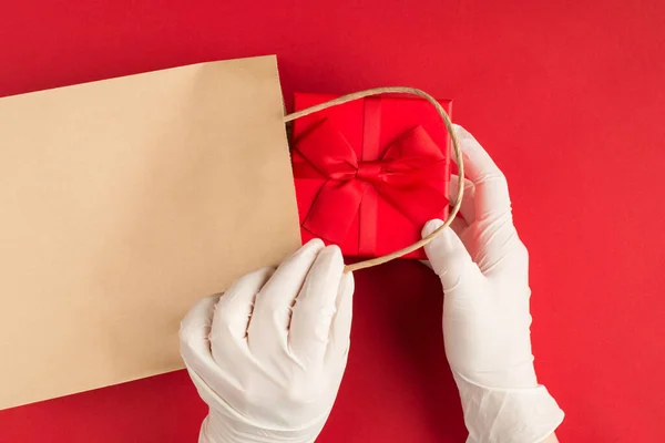 Presents give away during pandemic concept. Top above overhead view photo of female hands in gloves packing a red giftbox with red ribbon into craft papper bag isolated on red background