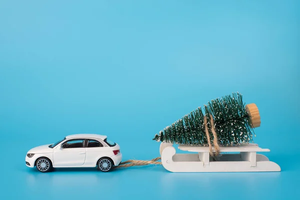 Christmas is coming soon concept. Close up photo of toy mini car carrying little christmas tree on white wooden sledge isolated on blue background