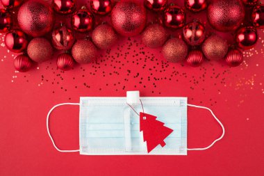 Noel ve karantina konsepti. Üst tepede güzel dekore edilmiş kırmızı takılar konfeti maskesi ve el dezenfektanı kırmızı arka planda izole edilmiş noel ağacı şeklindeki etiketli.