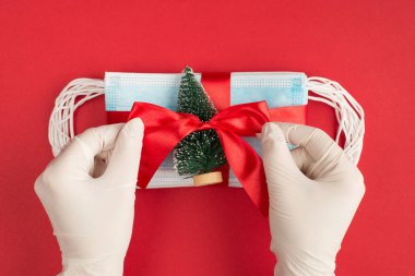 Tepede, beyaz lateks eldivenli ellerin Noel hediyesini sarmaladığı parlak kırmızı arka planda izole edilmiş bir fotoğraf var.
